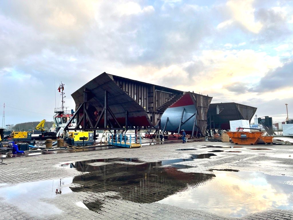 Große Stahlsektionen des Transportschiffs Asuka III werden auf einer Werft gebaut, mit Ausrüstung, Arbeitern und einem bewölkten Himmel, der sich im Wasser auf dem Boden spiegelt.
