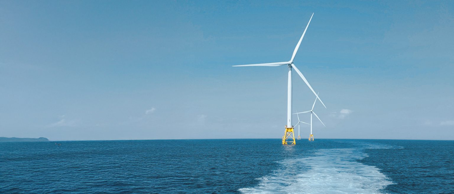 Three offshore wind turbines stand in the ocean under a clear blue sky, with a boat wake visible in the water leading towards them.