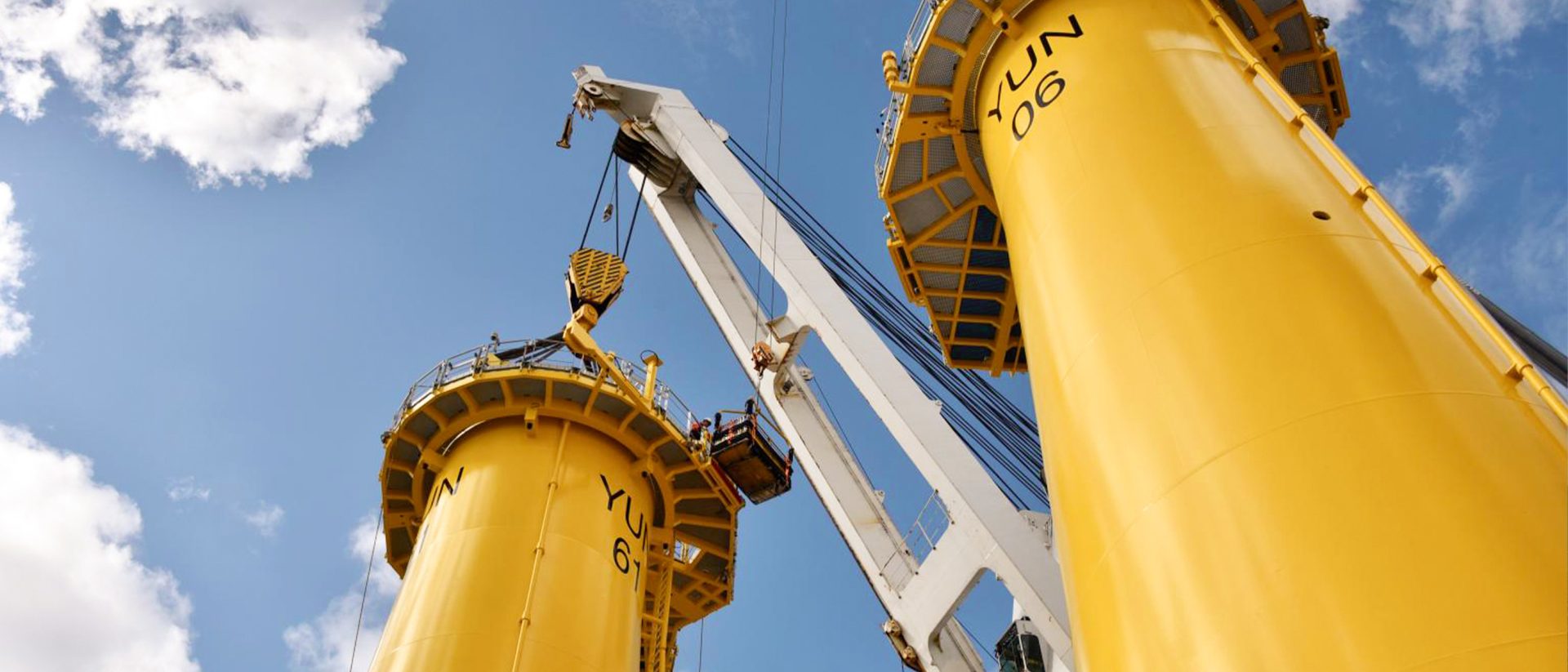 Close-up of a crane lifting equipment between two large yellow industrial structures labeled "YUN 06" and "YUN 61" against a partly cloudy sky.