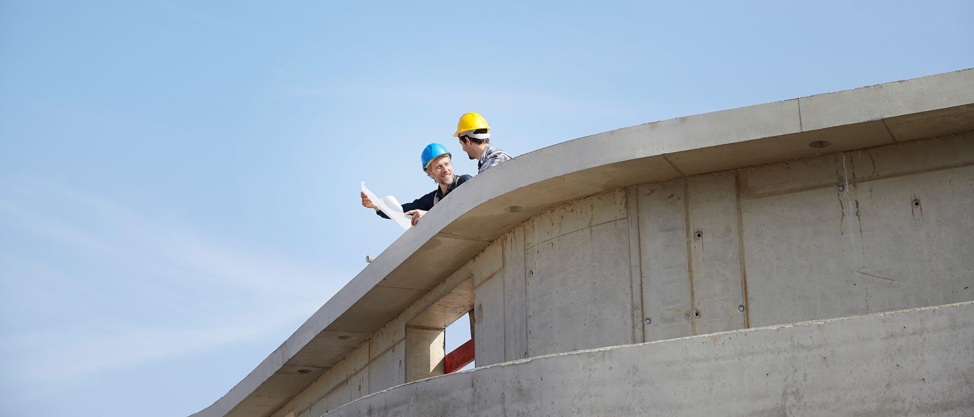 Zwei Bauarbeiter mit Helmen stehen auf einer Betonkonstruktion und prüfen unter einem klaren blauen Himmel Baupläne für ein Hoch- und Ingenieurbauprojekt.