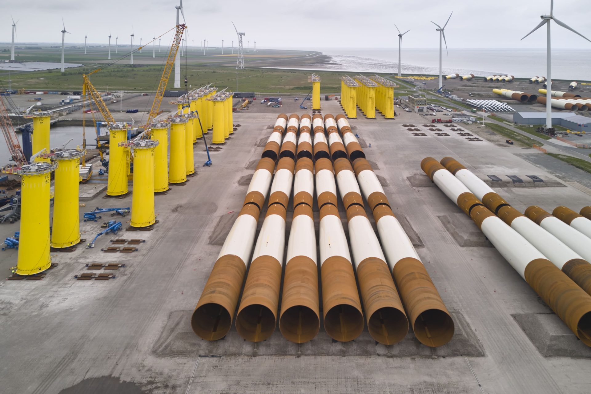 Large cylindrical steel segments and yellow foundation structures for offshore wind turbines are arranged on an industrial site near the coast, with wind turbines visible in the background.