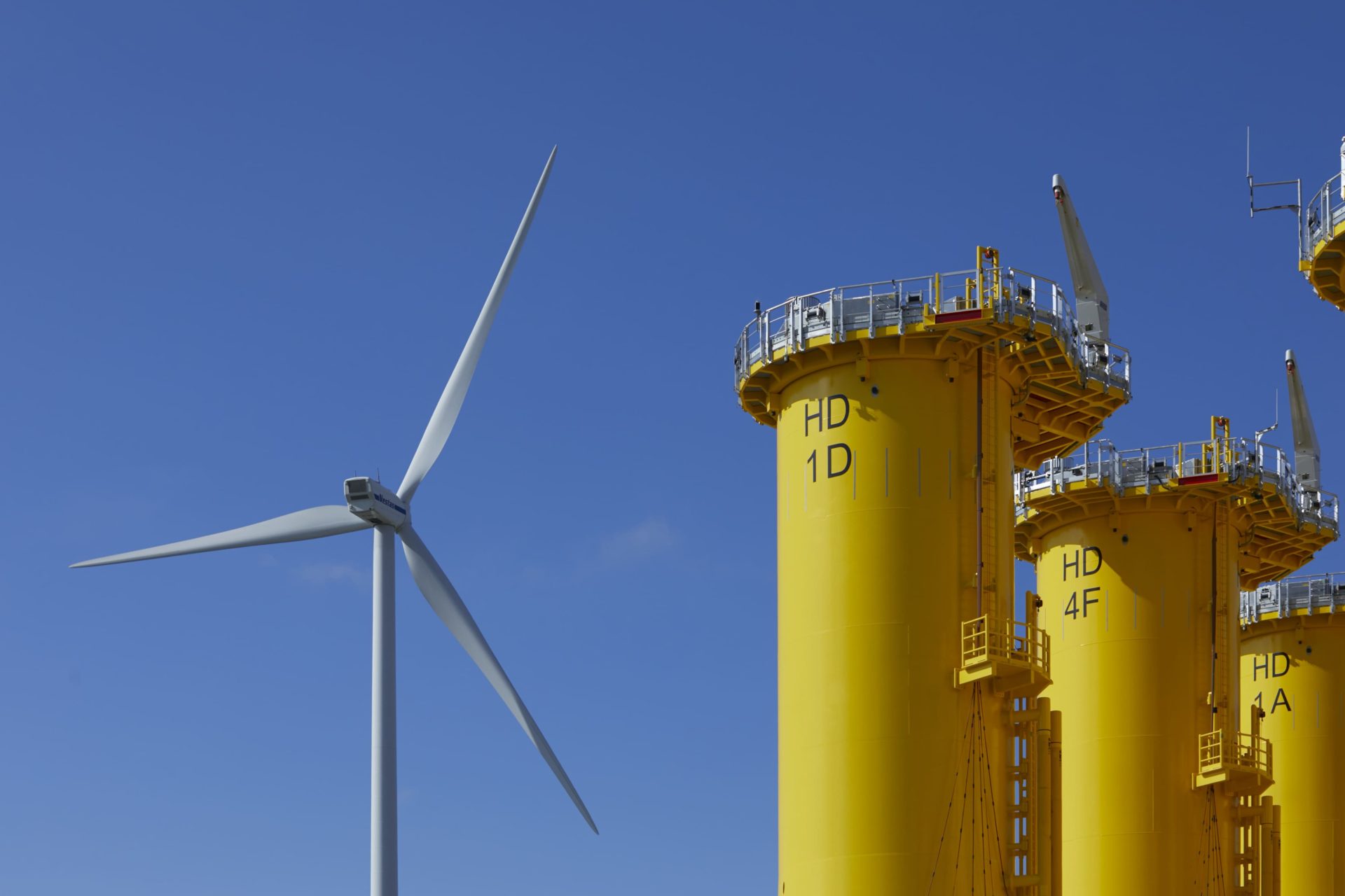 A wind turbine stands next to several large yellow industrial structures marked with codes under a clear blue sky.