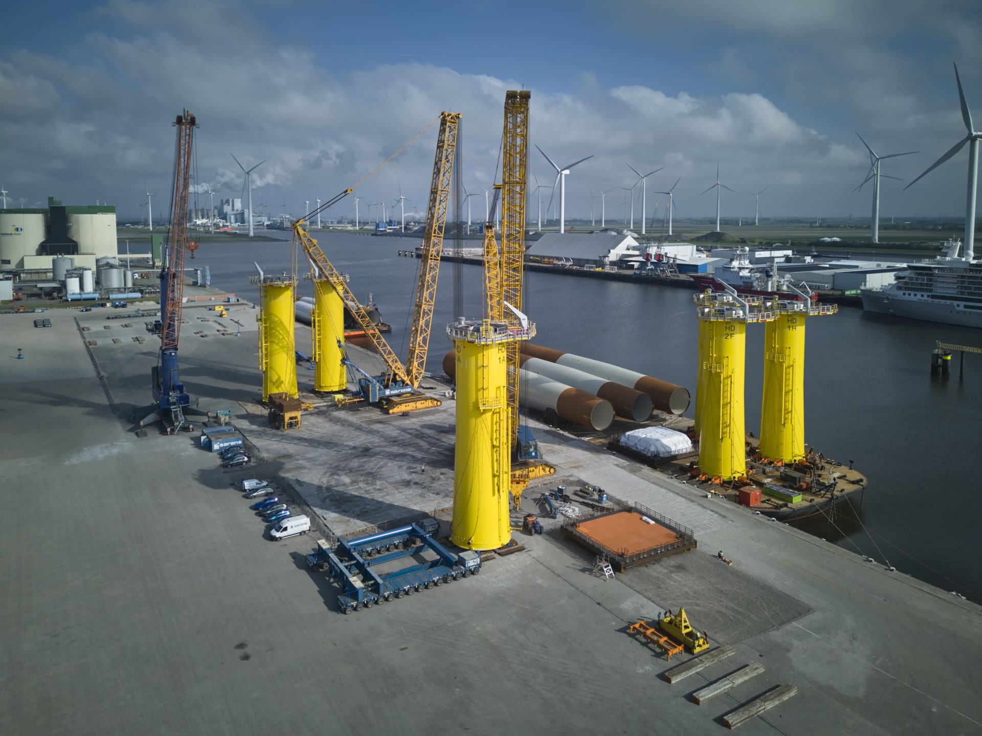 A dockyard scene with cranes assembling large yellow offshore wind turbine foundations, wind turbines and industrial buildings visible in the background.