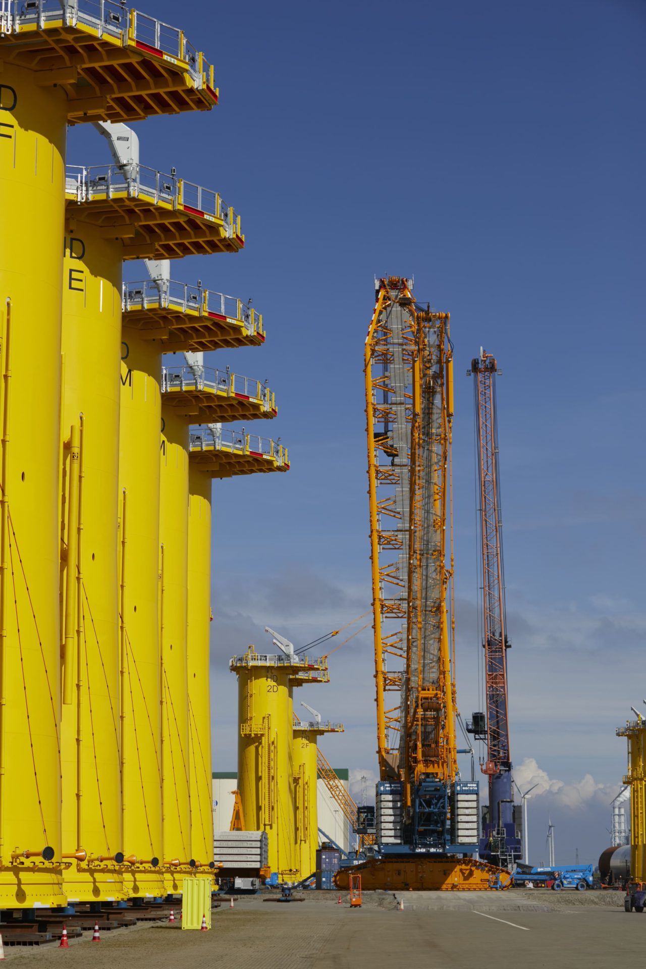 Large yellow offshore wind turbine foundations stand upright near tall cranes at an industrial dock under a clear blue sky.
