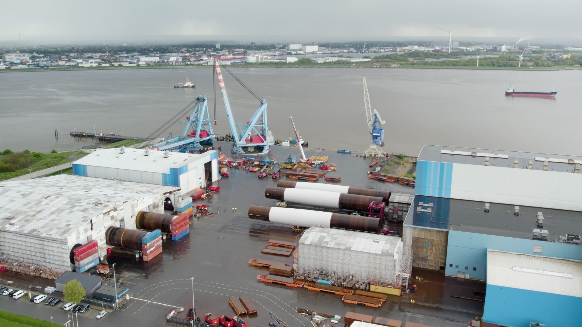 Aerial view of an industrial port with cranes, large cylindrical metal structures, warehouses, and ships on a calm river under a cloudy sky.