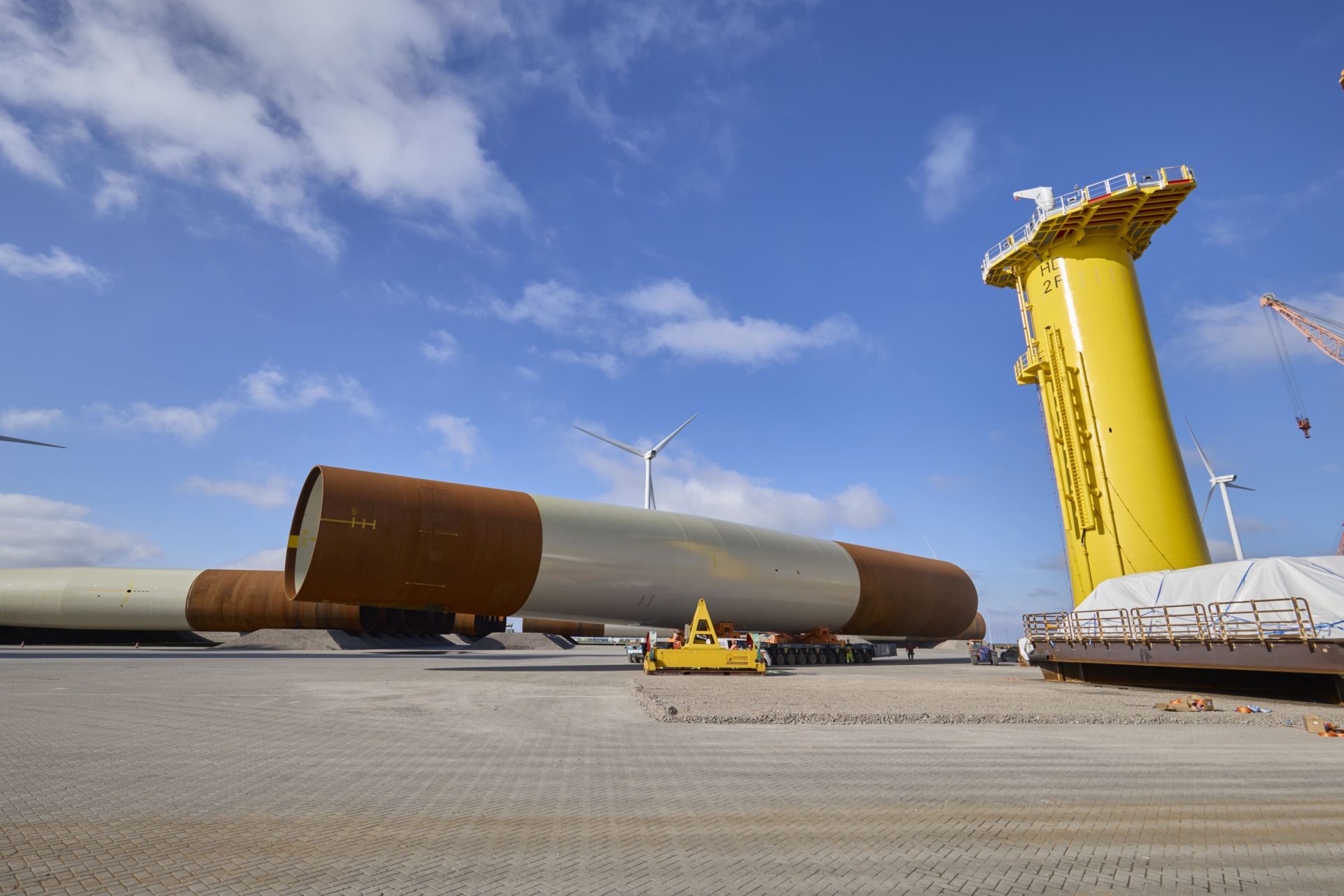 Several large cylindrical steel wind turbine foundations and a tall yellow structure are positioned on a paved industrial site under a partly cloudy sky, with wind turbines in the background.