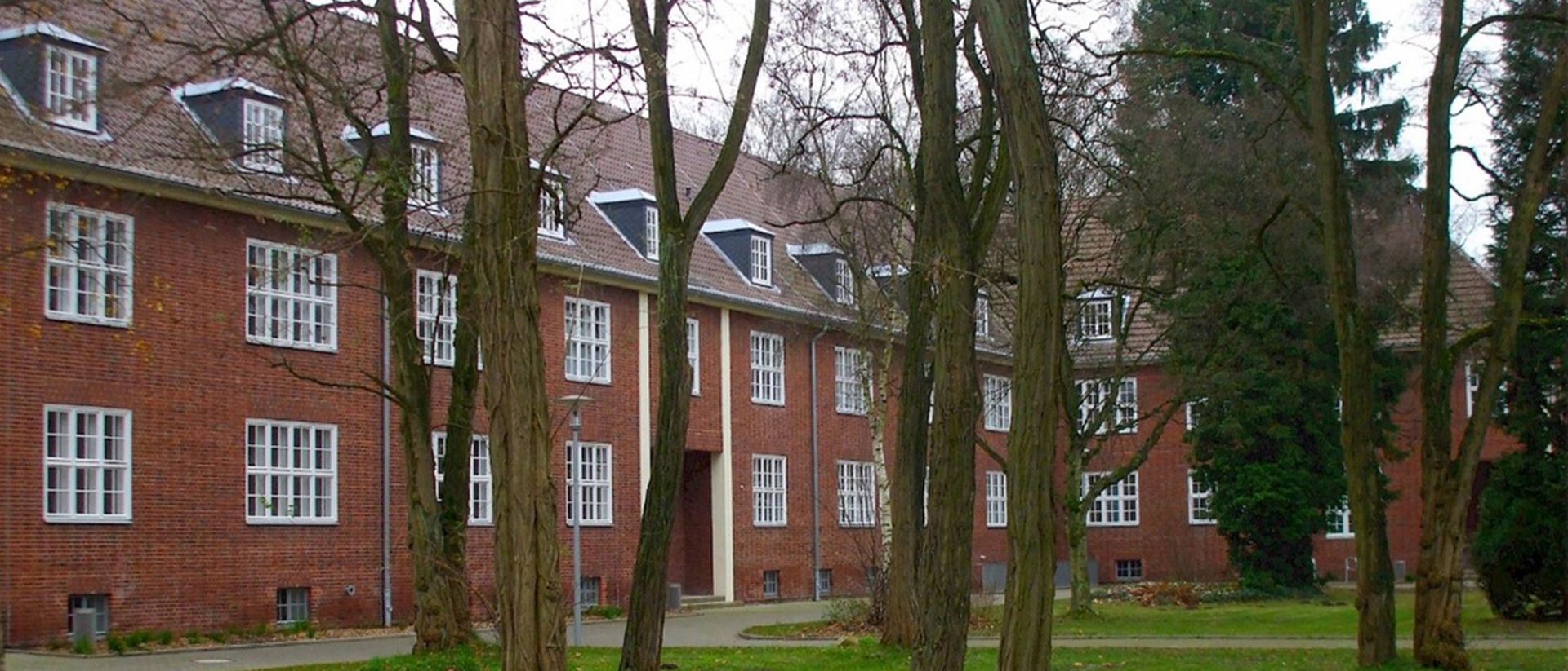 A red brick building with many white-framed windows, dormer windows on a sloped roof, and several leafless trees in the foreground.