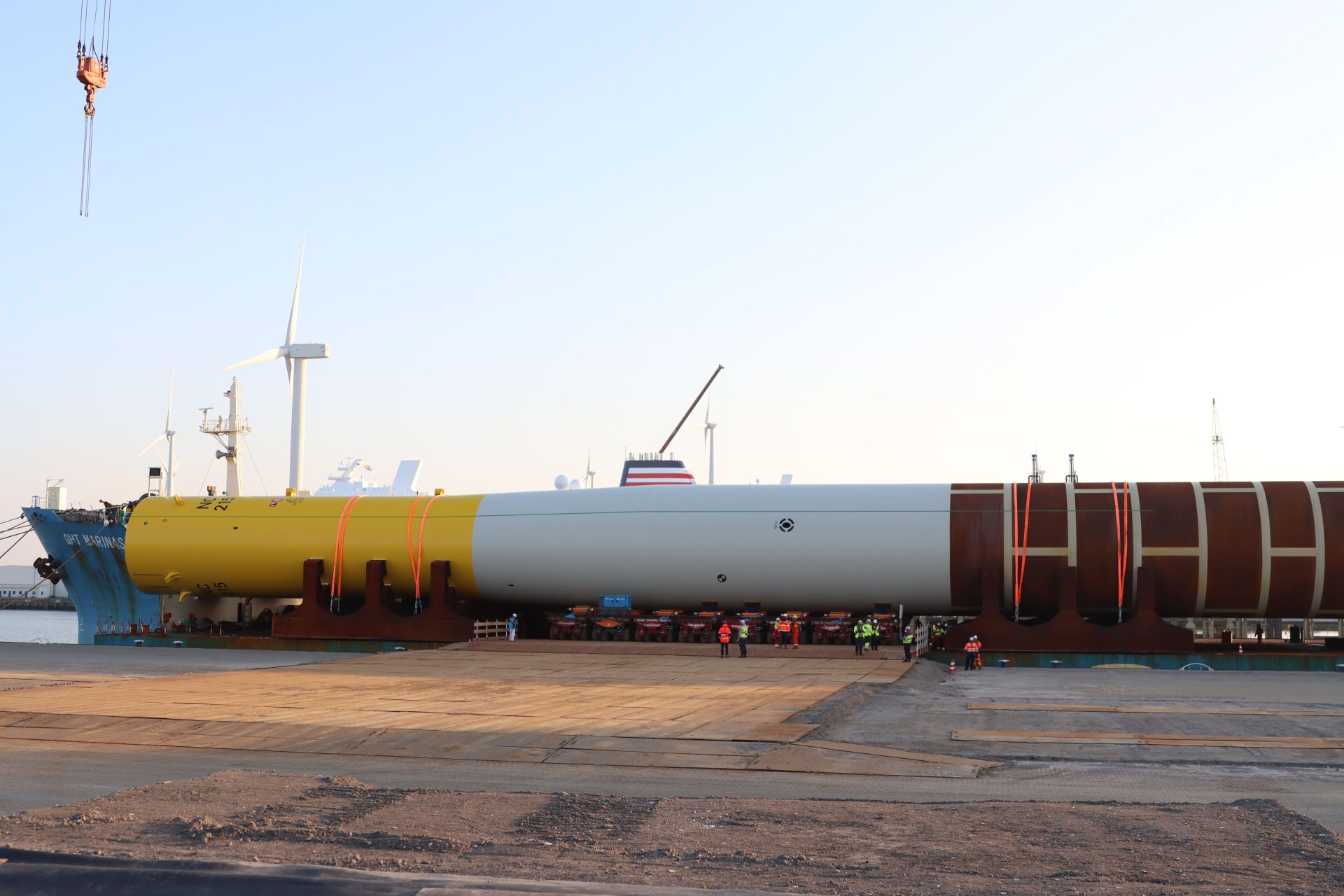 A large cylindrical wind turbine tower section is positioned horizontally on supports at an industrial port, with workers and cranes nearby. Wind turbines and cranes are in the background.