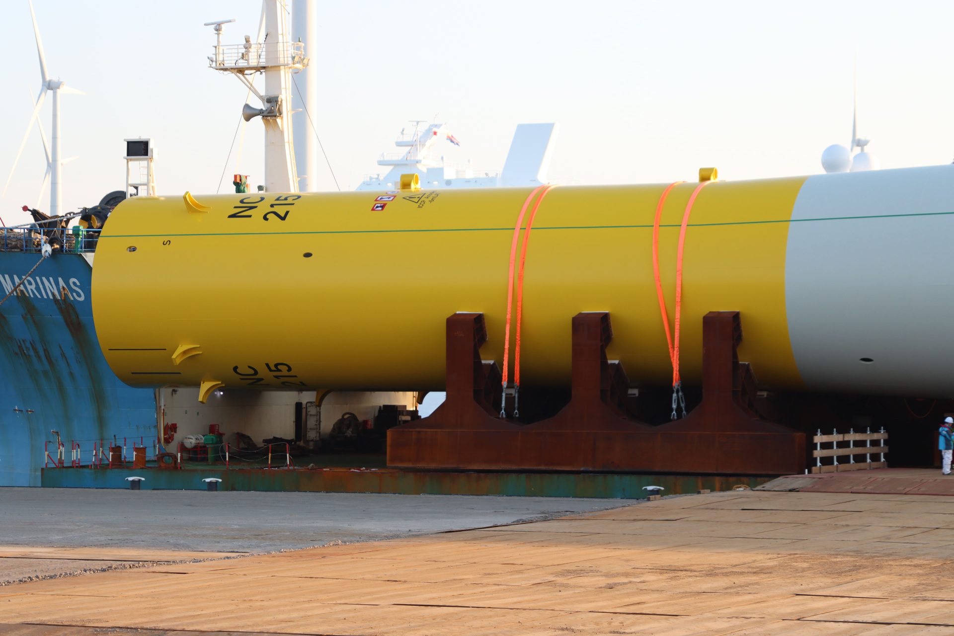A large yellow cylindrical industrial structure secured with orange straps rests on a dock, with a ship and wind turbines visible in the background.