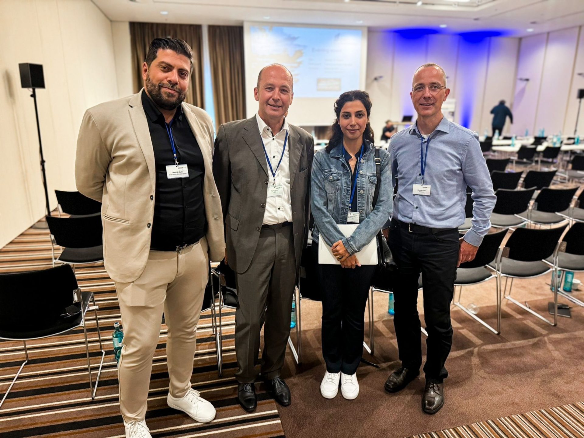 Four people stand together in a conference room at the Offshore Wind Event 2024 Bremen JBO, all wearing name badges, with rows of chairs and a presentation screen in the background.