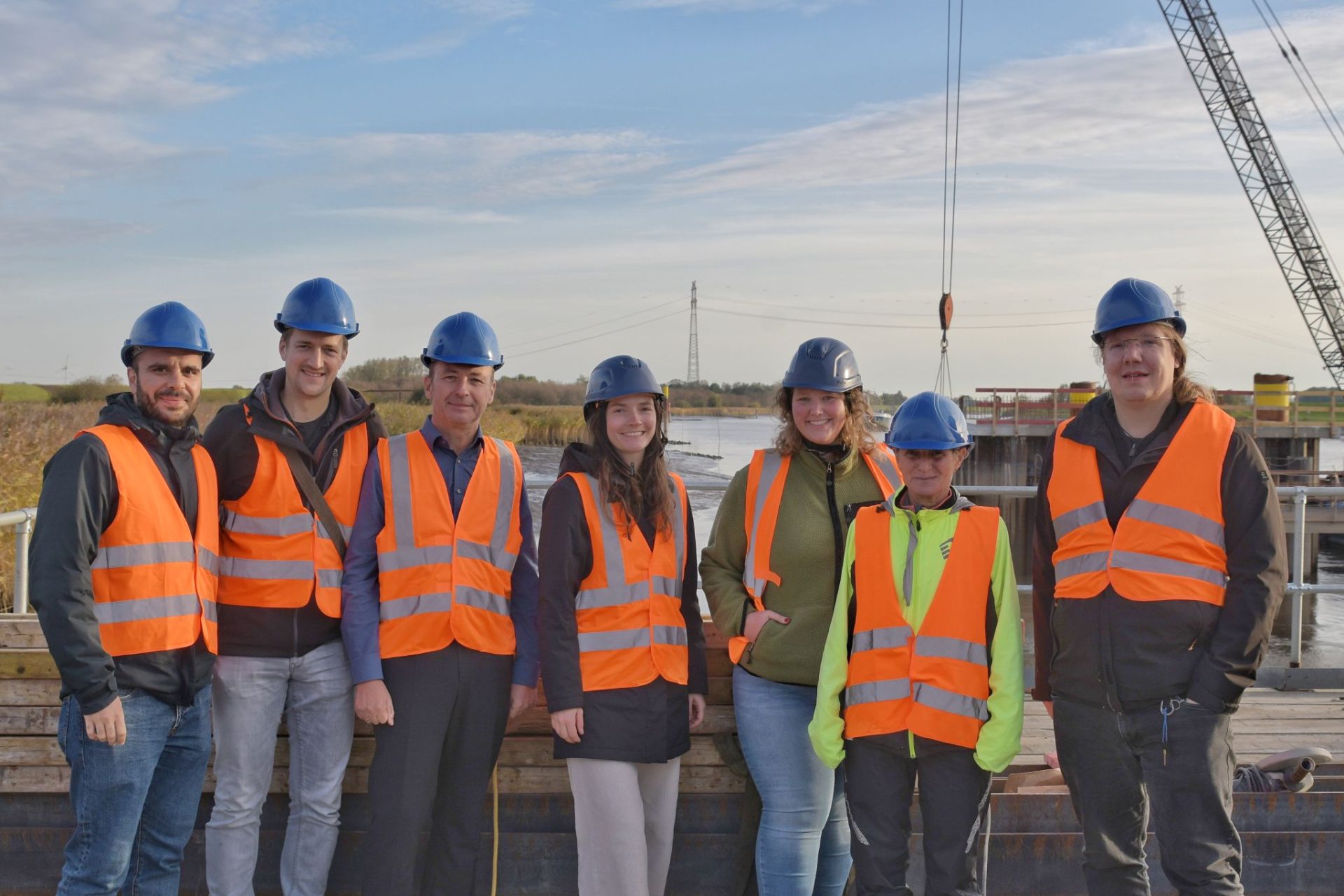 Seven people wearing blue hard hats and orange safety vests stand together at the Friesenbrücke Deutsche Bahn construction site near a body of water, with construction equipment in the background.