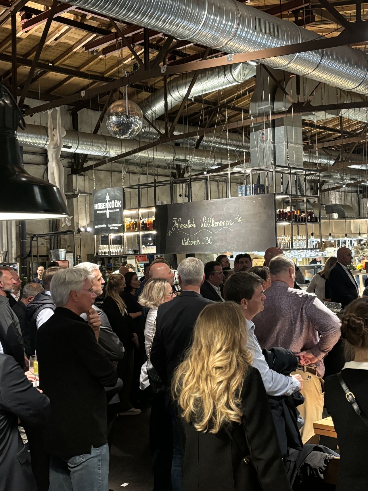 A crowd of people stands and listens in an industrial-style indoor venue with exposed pipes, hanging lights, and a chalkboard sign behind the bar area, celebrating 10 years of JBO Offshore WindEnergy Hamburg.