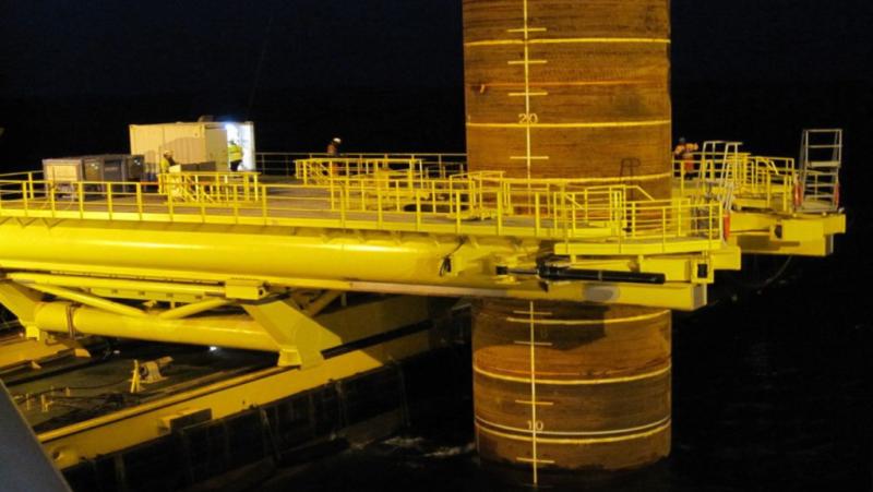A large yellow offshore platform structure is attached to a cylindrical column marked with measurement lines, with workers and equipment visible on the platform at night.