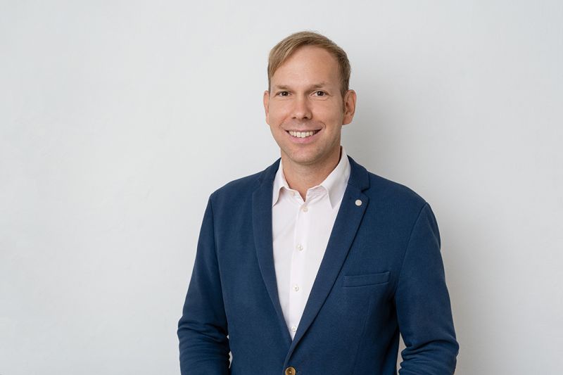 A man wearing a blue blazer and white shirt stands in front of a plain light background, smiling at the camera.
