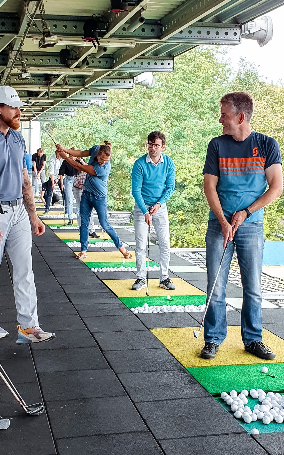 Several people stand on individual mats at a driving range, holding golf clubs and practicing their swings, with baskets of golf balls nearby.