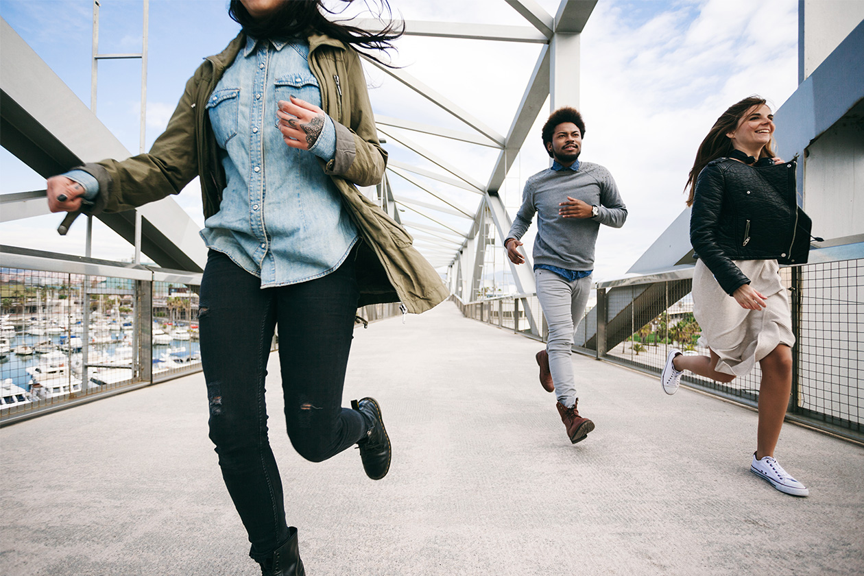 Drei Menschen, zwei Frauen und ein Mann, laufen und lächeln über eine moderne Fußgängerbrücke, im Hintergrund ist ein Jachthafen zu sehen.