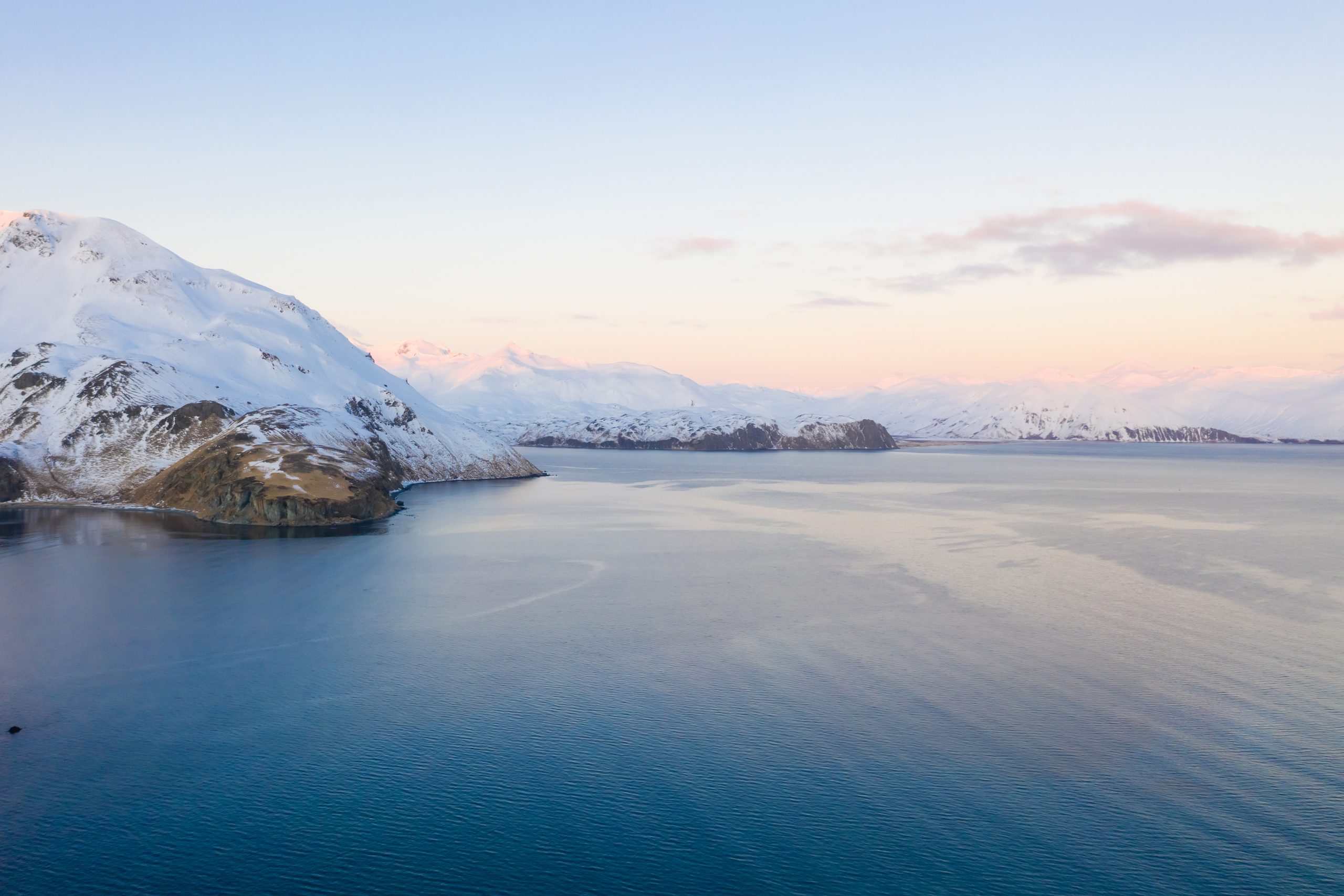 Ein ruhiges Gewässer mit schneebedeckten Bergen im Hintergrund unter einem pastellfarbenen Himmel bei Sonnenuntergang.