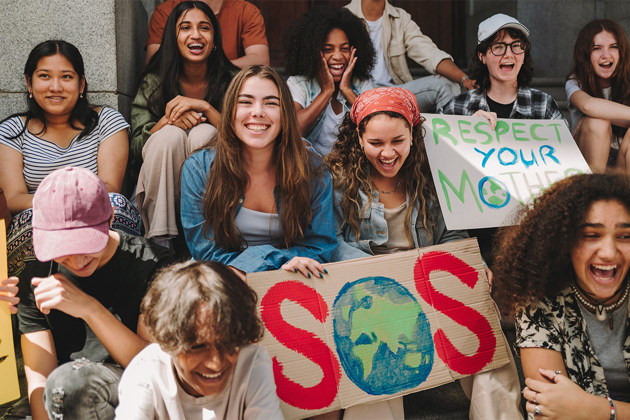 Eine bunt gemischte Gruppe junger Leute sitzt auf Stufen, einige halten Schilder mit der Aufschrift „SOS mit einem Globus“ und „Respektiere deine Mutter“ hoch und nehmen offenbar an einem Klimaprotest teil.