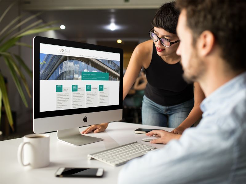 Two people look at a computer screen displaying a corporate website, with one person seated at the desk and the other standing and pointing at the monitor.