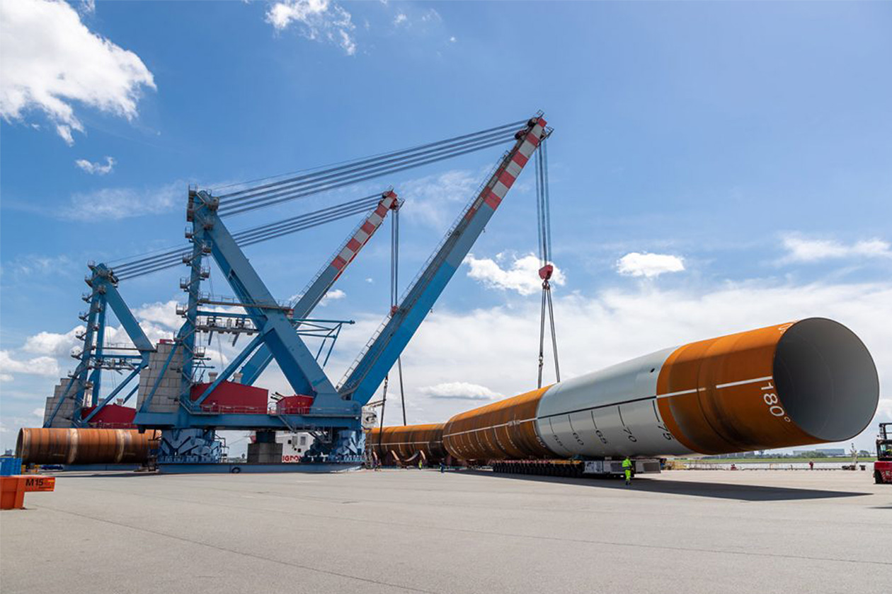 Große blaue Kräne heben und transportieren unter einem teilweise bewölkten Himmel ein massives orangefarbenes zylindrisches Rohr in einem Industriehafen.