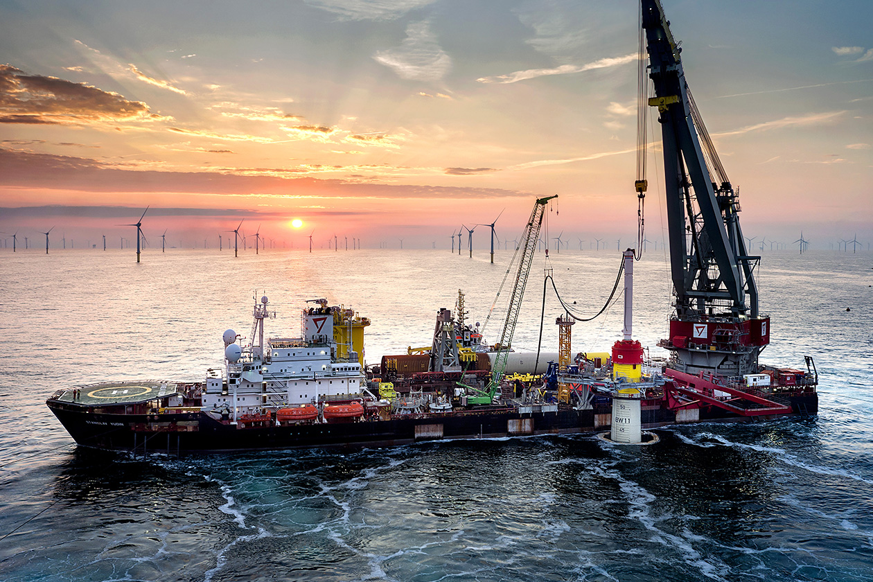 Ein großes Kranschiff installiert bei Sonnenuntergang auf See Ausrüstung. Im Hintergrund sind Windturbinen zu sehen, und das Schiff ist von ruhigem Wasser umgeben.