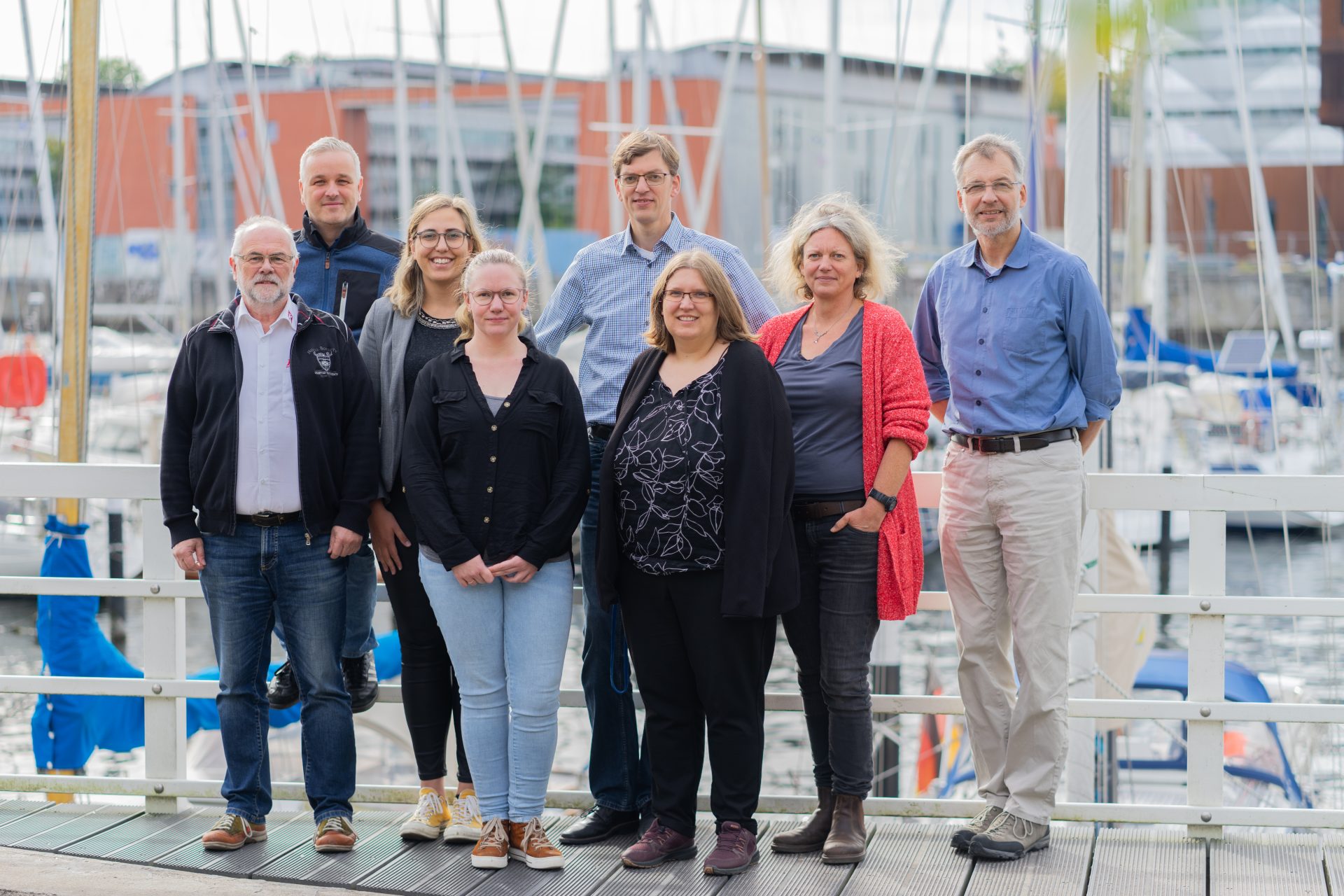 Eight adults stand outdoors in front of a marina with sailboats, posing for a group photo. They are dressed in casual clothing and appear to be smiling.
