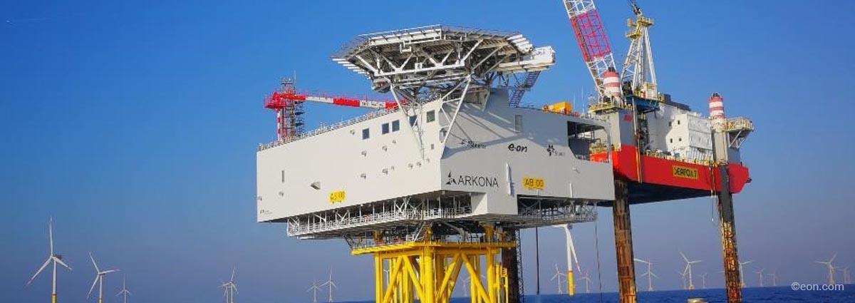 An offshore wind farm substation labeled "ARKONA" stands on yellow supports in the sea, surrounded by wind turbines and construction cranes against a clear blue sky.