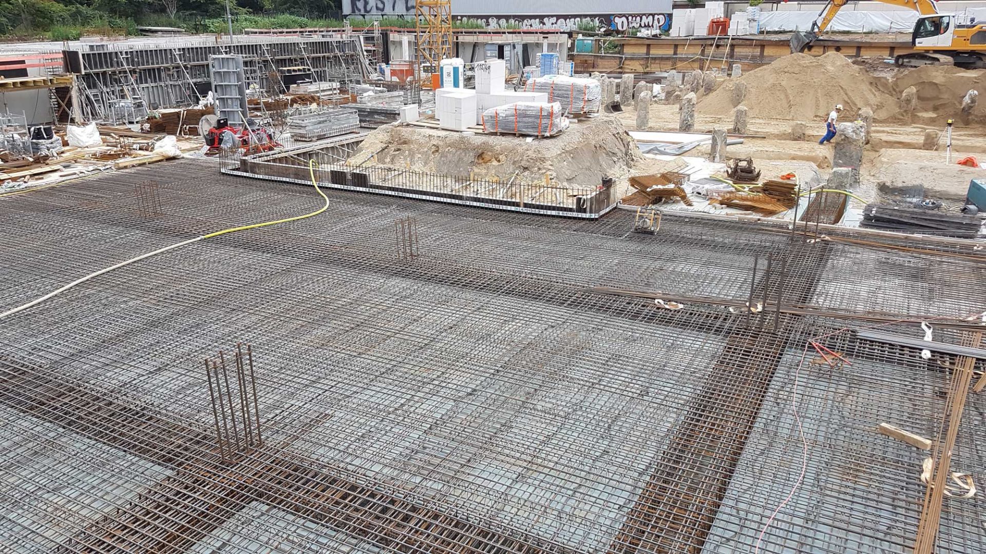 A construction site with steel reinforcement bars laid out for a concrete foundation, building materials, and workers visible in the background.