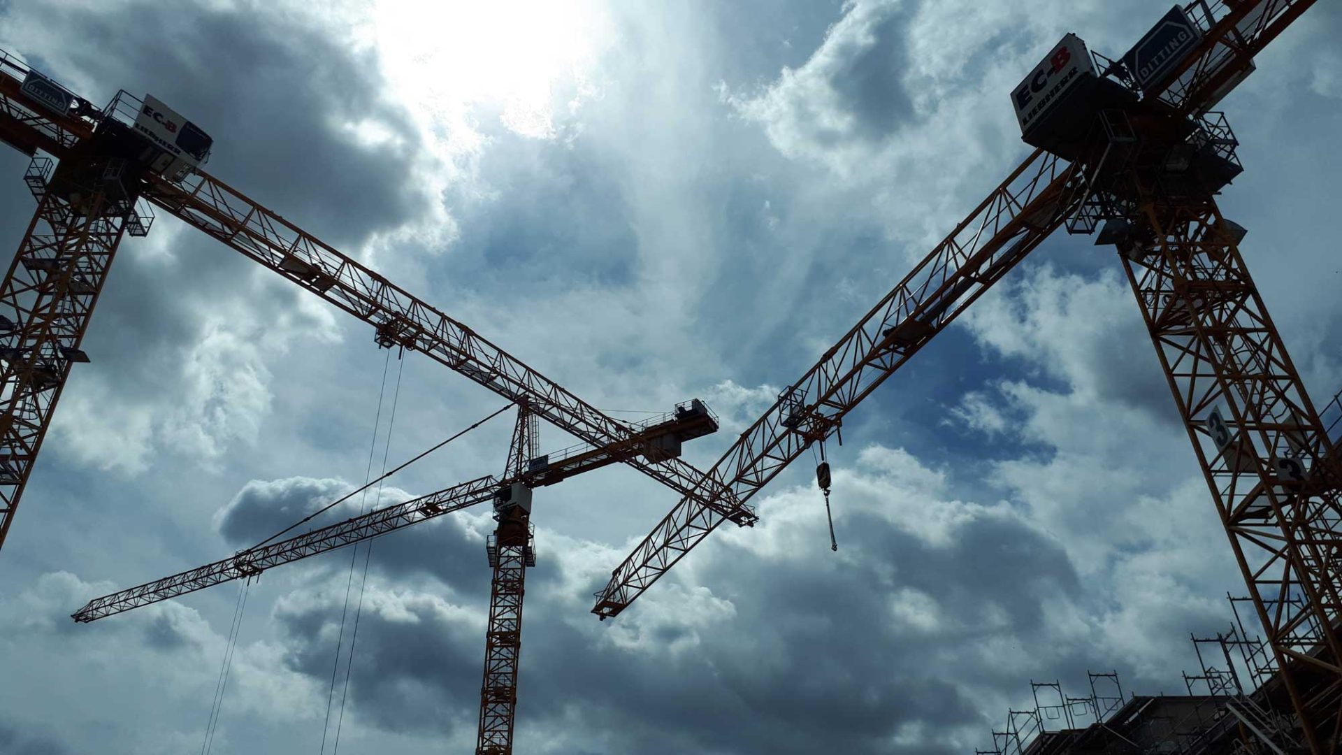 Four large construction cranes tower against a cloudy sky, with the sun partially obscured by clouds.