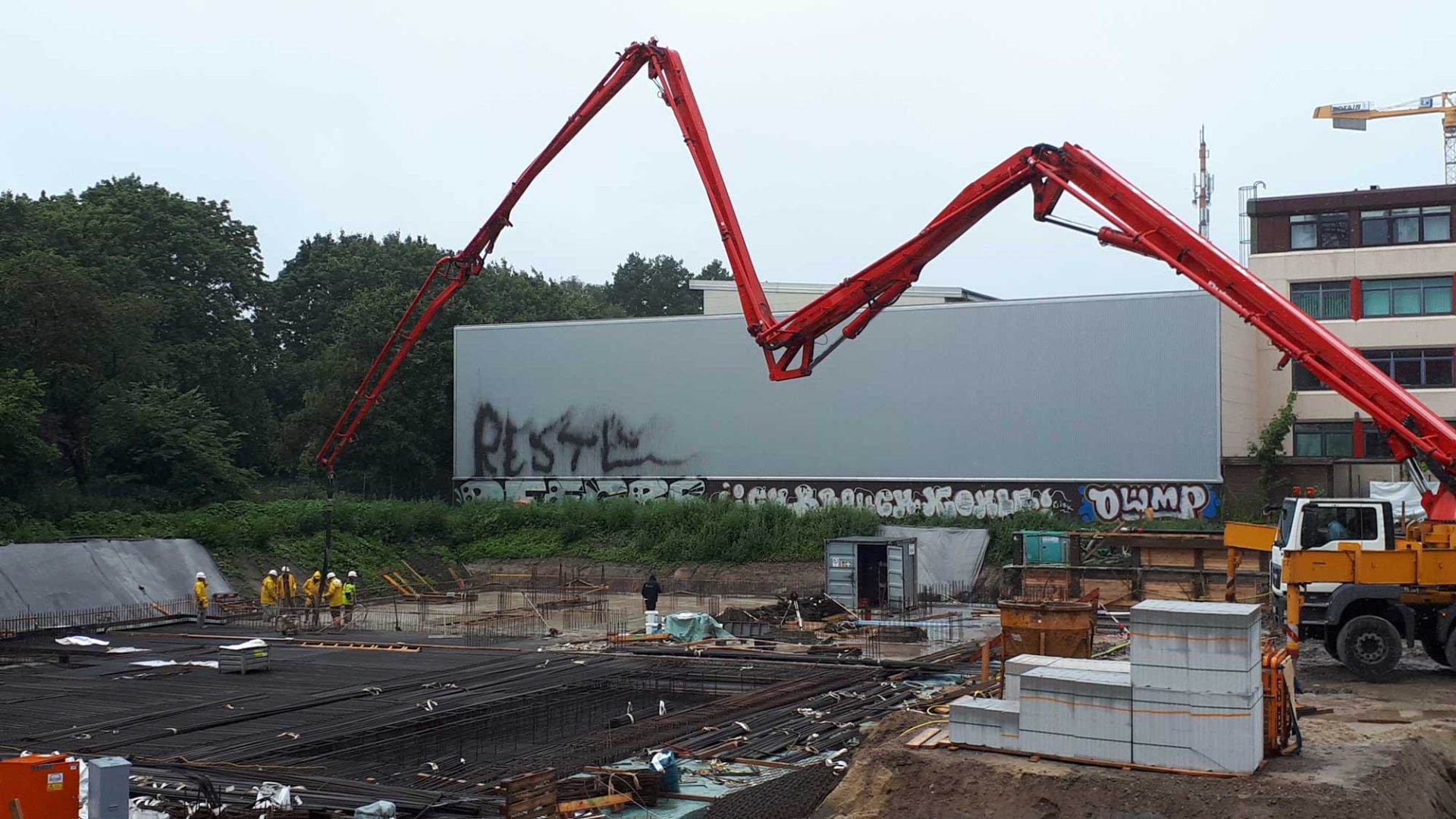Construction site with workers and a large concrete pump truck, steel reinforcements, and building materials; graffiti on a wall in the background.