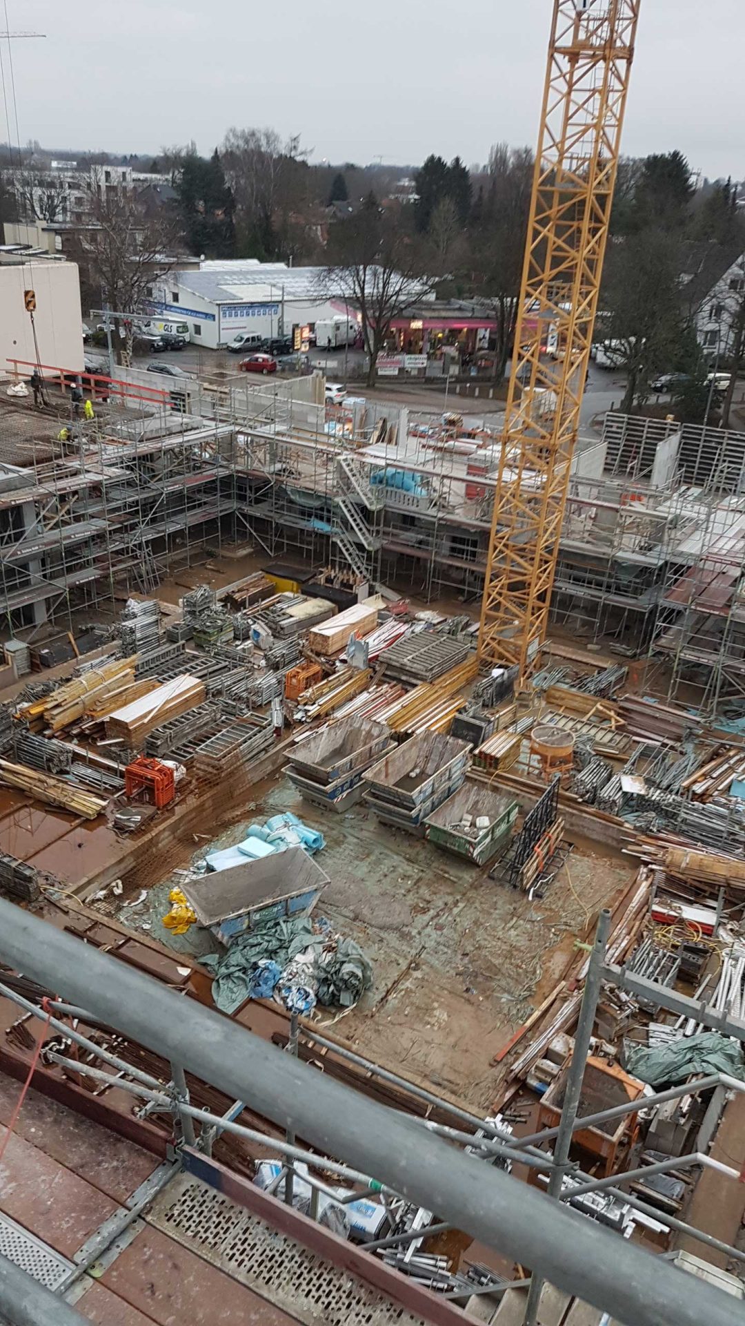 A construction site with scaffolding, building materials, and a yellow tower crane; surrounding trees and buildings visible in the background.