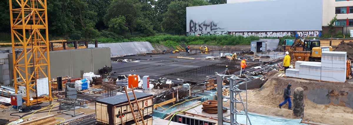 Baustelle mit Arbeitern, Maschinen und Baumaterialien; sichtbar sind Fundamentarbeiten und Stahlbewehrungen, im Hintergrund ein Kran und Graffiti an einer Wand.