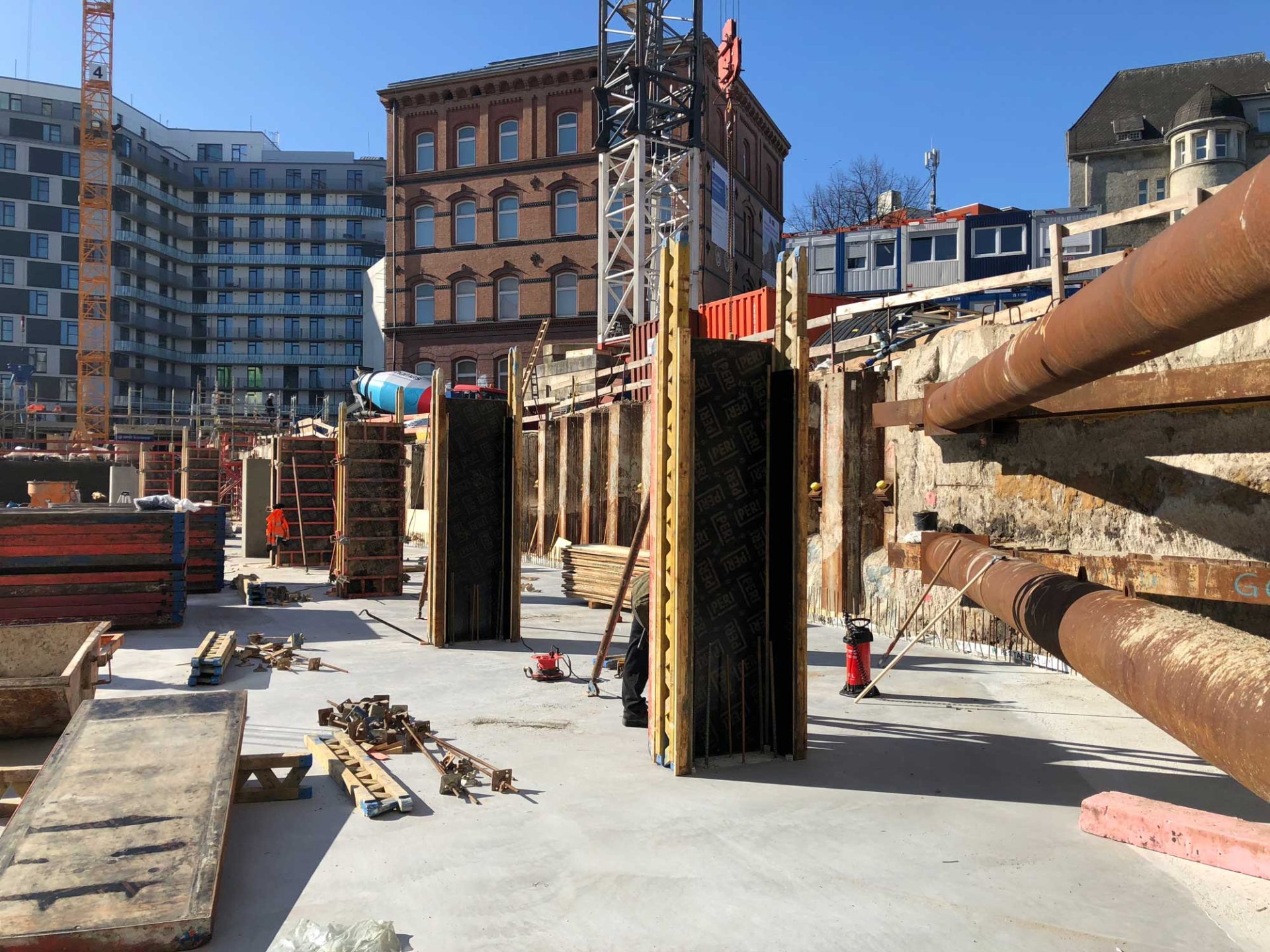 Baustelle mit Betonboden, Stahlträgern, Holzbrettern und Baumaterialien; im Hintergrund sind Kräne und mehrstöckige Gebäude zu sehen.