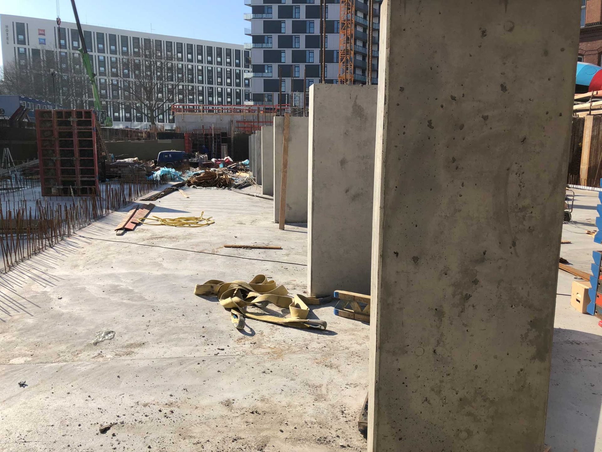 A construction site with concrete pillars, scattered materials, and rebar on the ground. Modern buildings are visible in the background.