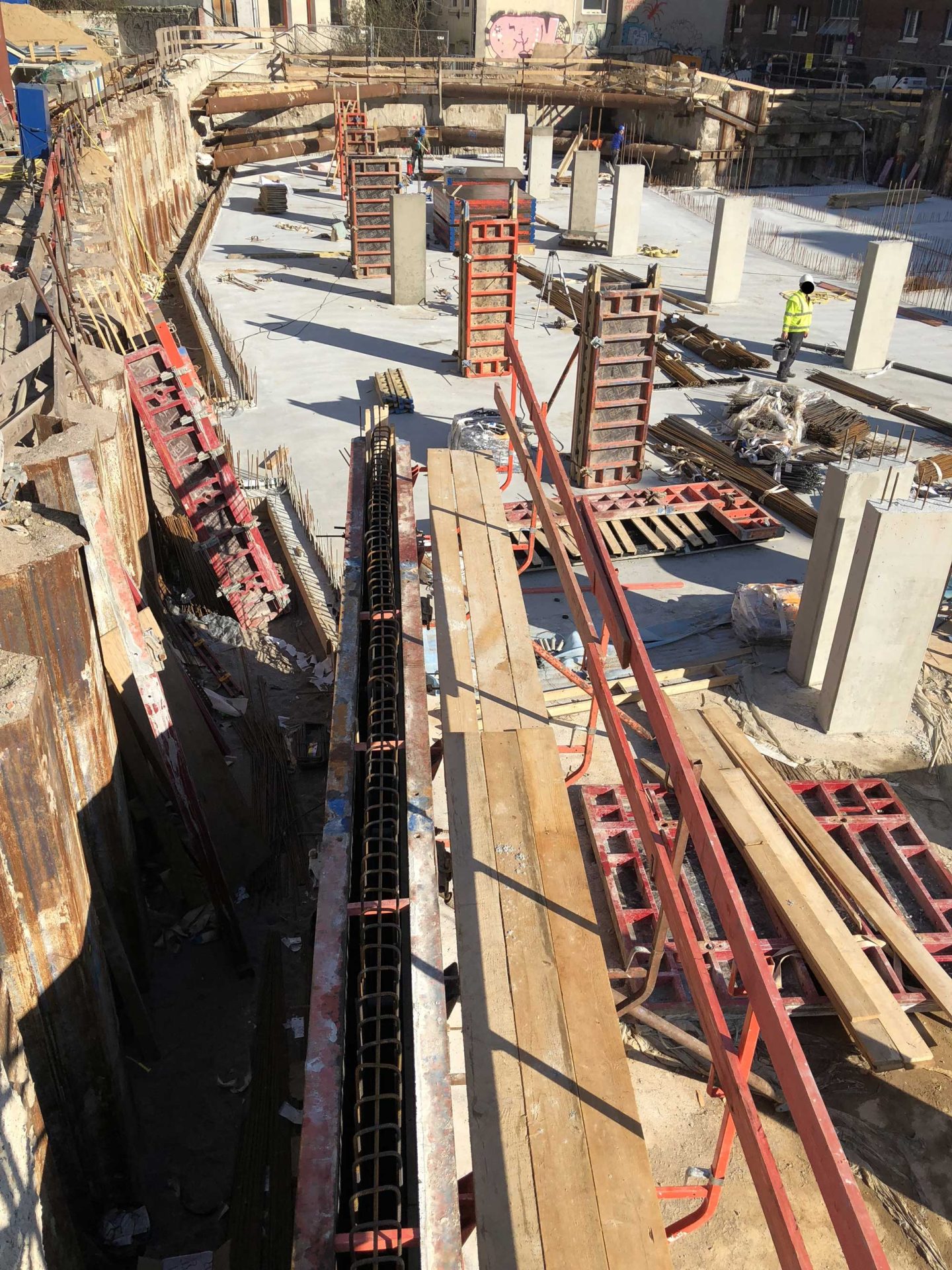 Construction site with concrete pillars, wooden planks, and metal scaffolding; a worker in a yellow vest stands in the background.