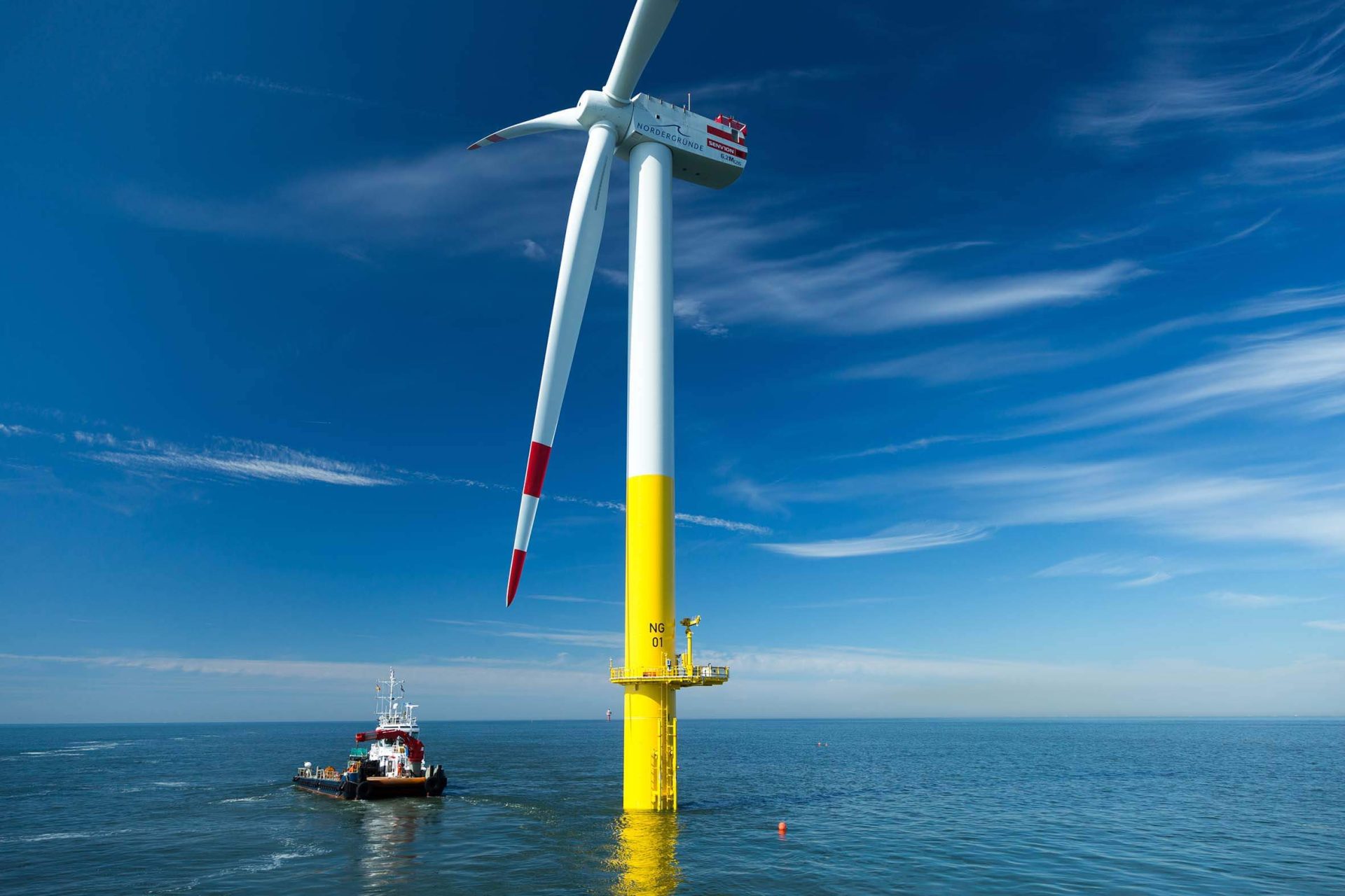 A large offshore wind turbine stands in the sea with a maintenance vessel nearby under a clear blue sky.