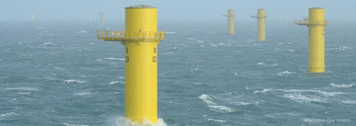 Yellow offshore wind turbine foundations rise from the sea on a cloudy day, with waves surrounding the structures and no turbine blades installed yet.