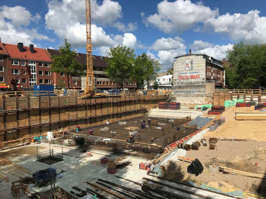 Construction site with workers installing rebar in a large foundation pit; a crane and surrounding buildings are visible under a partly cloudy sky.