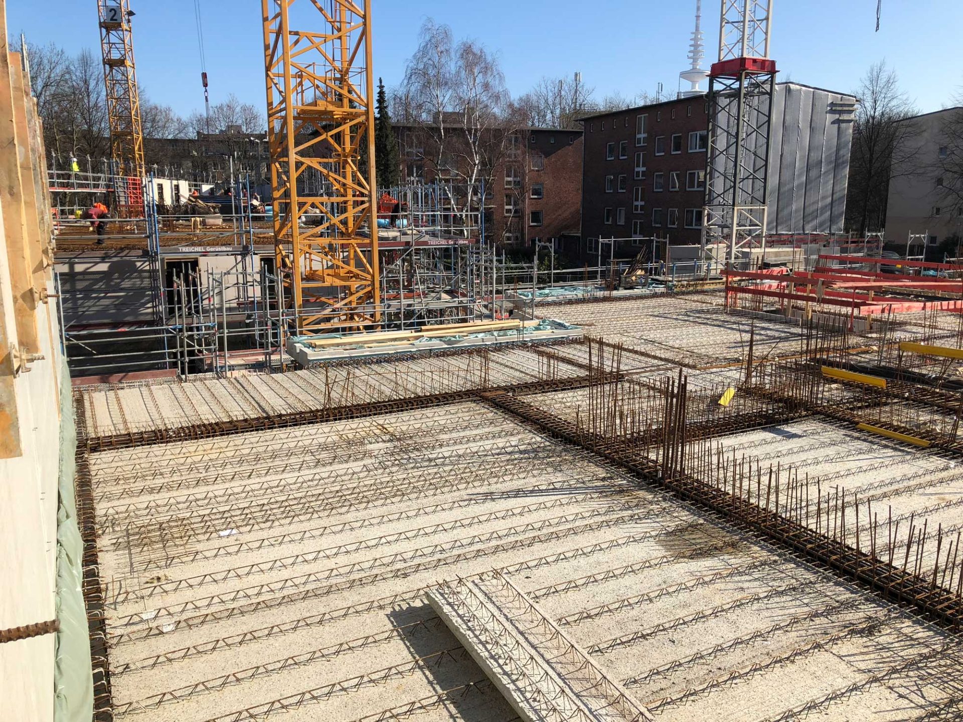 Construction site with exposed steel reinforcement bars, scaffolding, and cranes; buildings and trees are visible in the background under clear daylight.