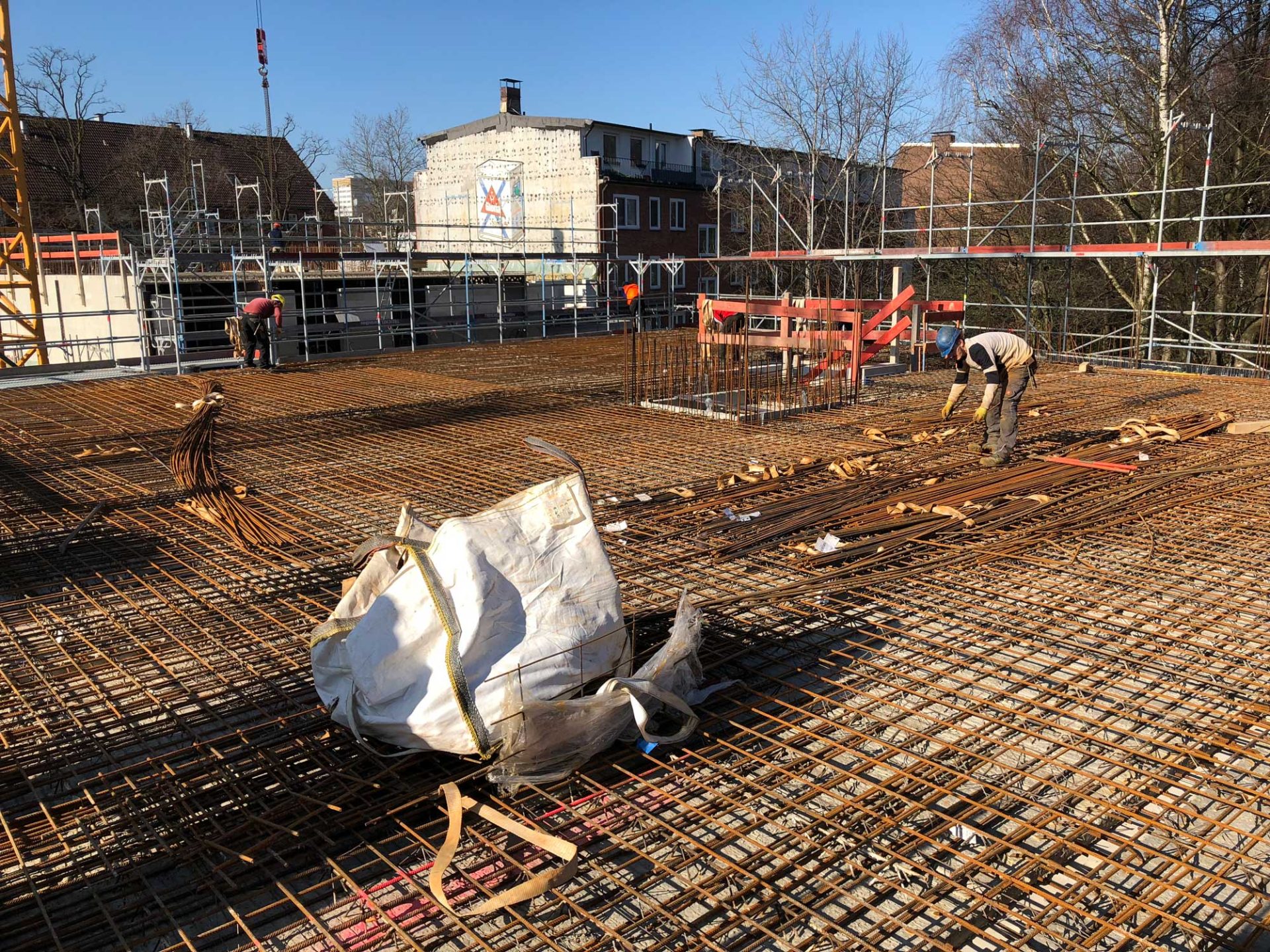 Construction workers arranging steel reinforcement bars on a large concrete slab at a building site, surrounded by scaffolding, with a white bag in the foreground.