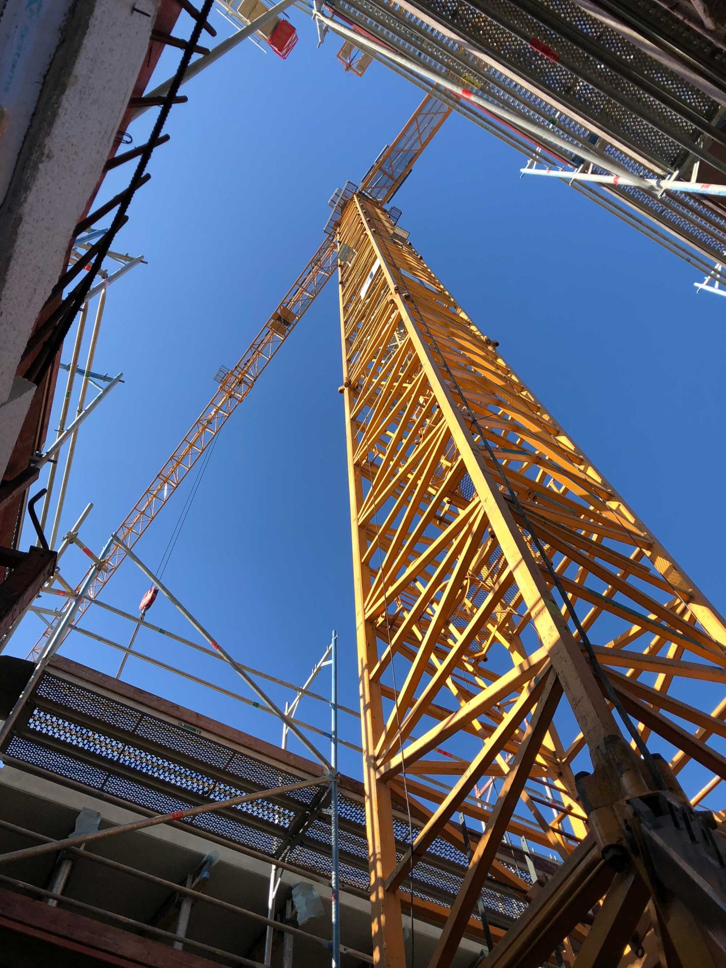 View looking up at a tall yellow construction crane surrounded by scaffolding and partially constructed buildings against a clear blue sky.