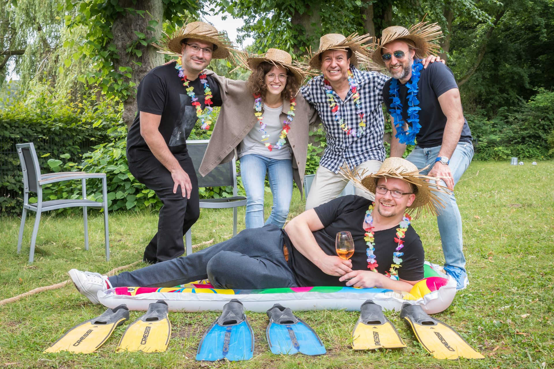 Six adults wearing straw hats and leis pose outdoors, with one person lying on an inflatable raft. Four pairs of swim fins are placed on the grass in front of them.