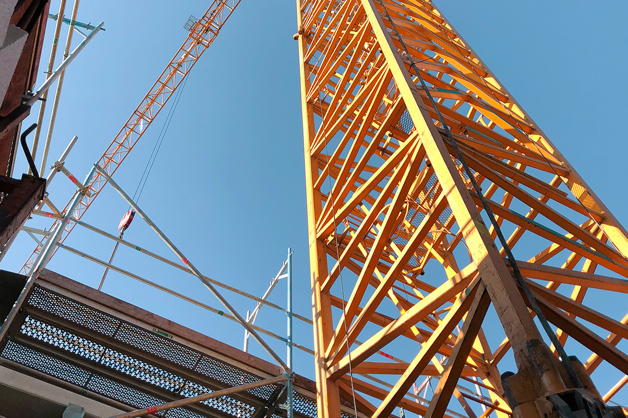 Blick von unten auf eine Baustelle mit gelben Kränen und Gerüsten vor einem klaren blauen Himmel.
