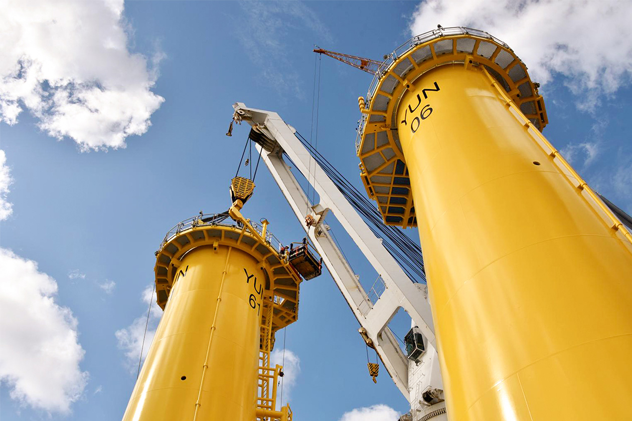 Zwei große gelbe Fundamente für Offshore-Windkraftanlagen werden vor einem teilweise bewölkten Himmel von einem Kran an ihren Platz gehoben.