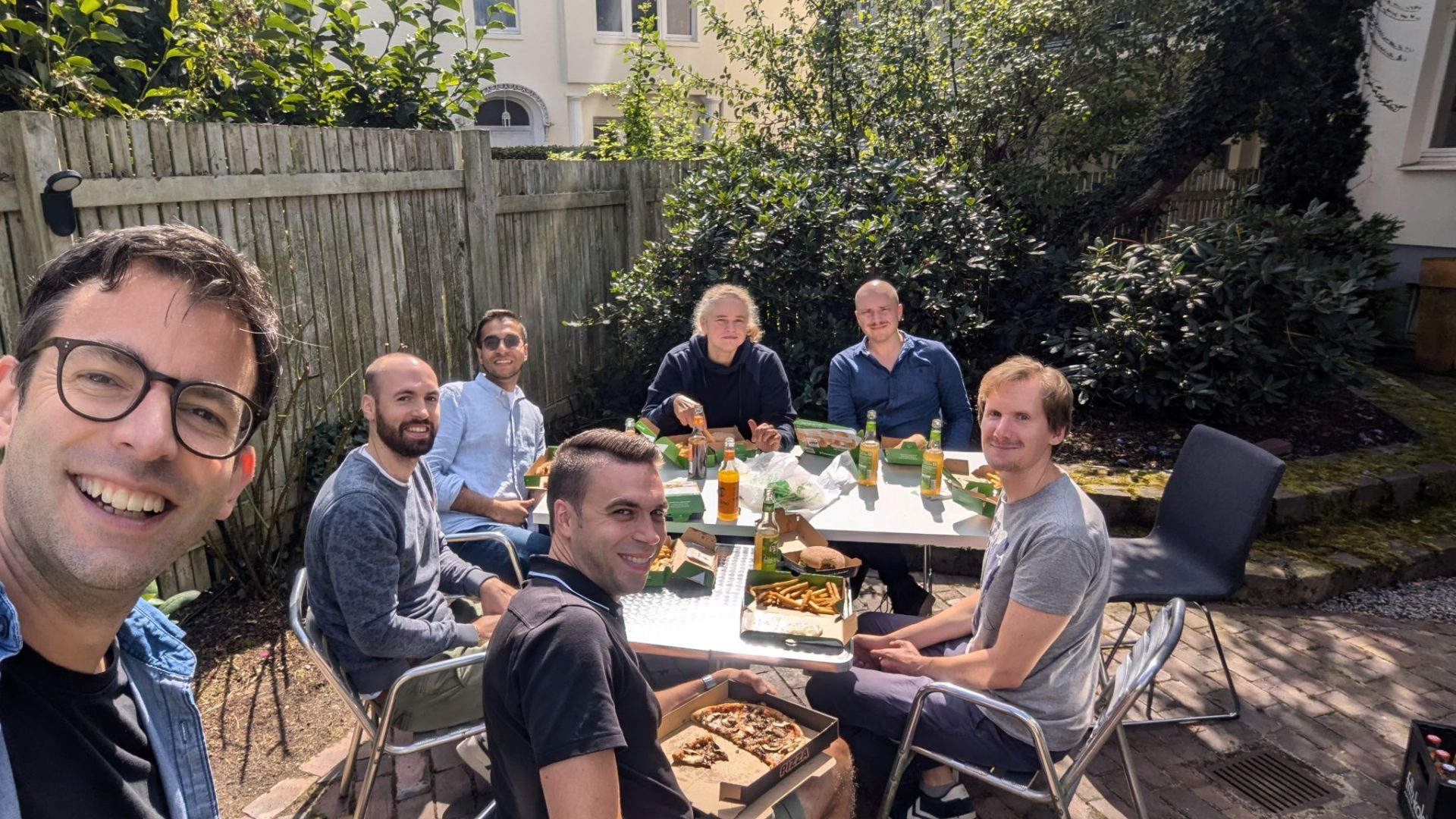 Seven people sit around a table outdoors eating pizza and fries, with drinks and food containers on the table, and greenery in the background.