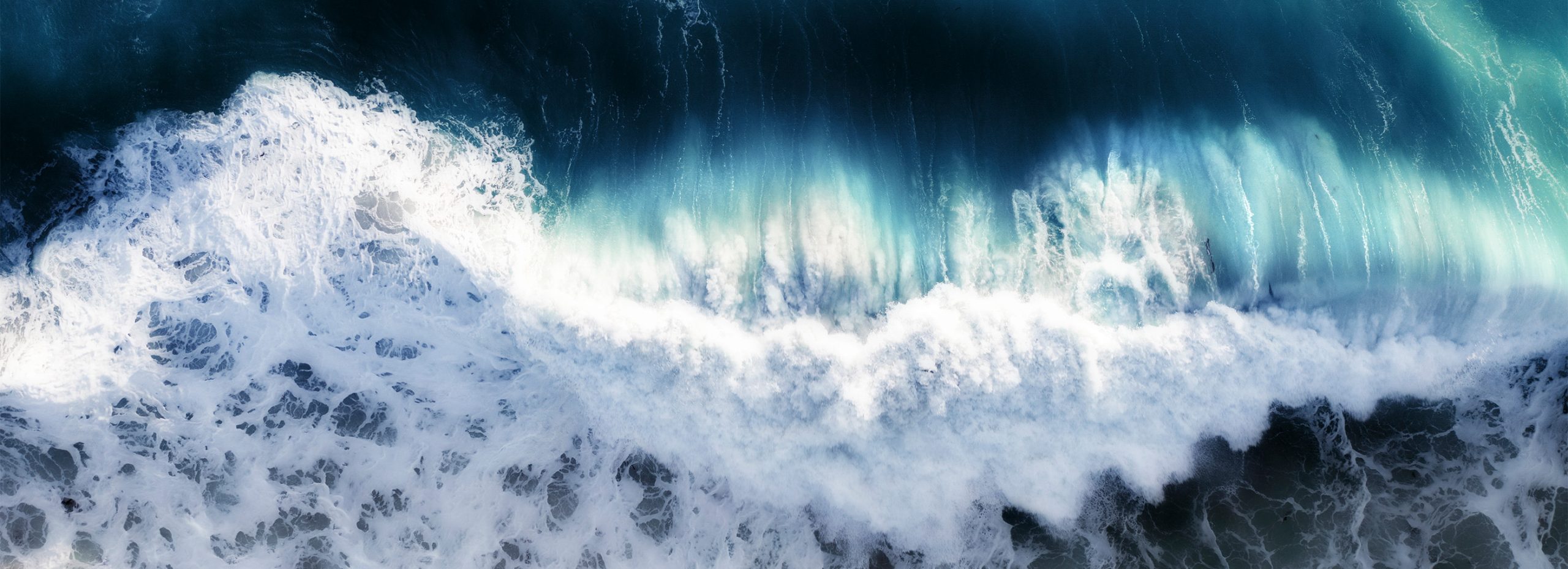 Luftaufnahme von Wellen, die an die Küste schlagen und weißen Schaum vor dem tiefblauen und türkisfarbenen Wasser bilden.