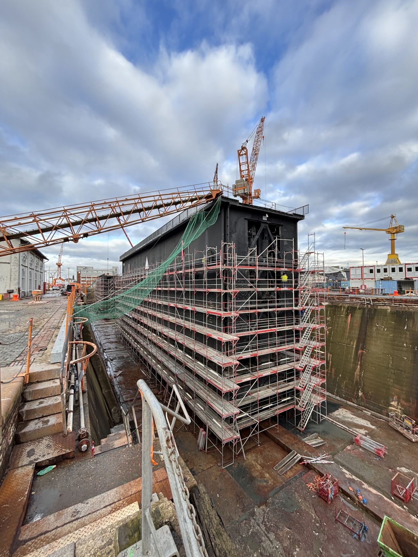 A large ship or structure is surrounded by scaffolding in a dry dock, with a crane overhead and construction materials scattered nearby under a cloudy sky.