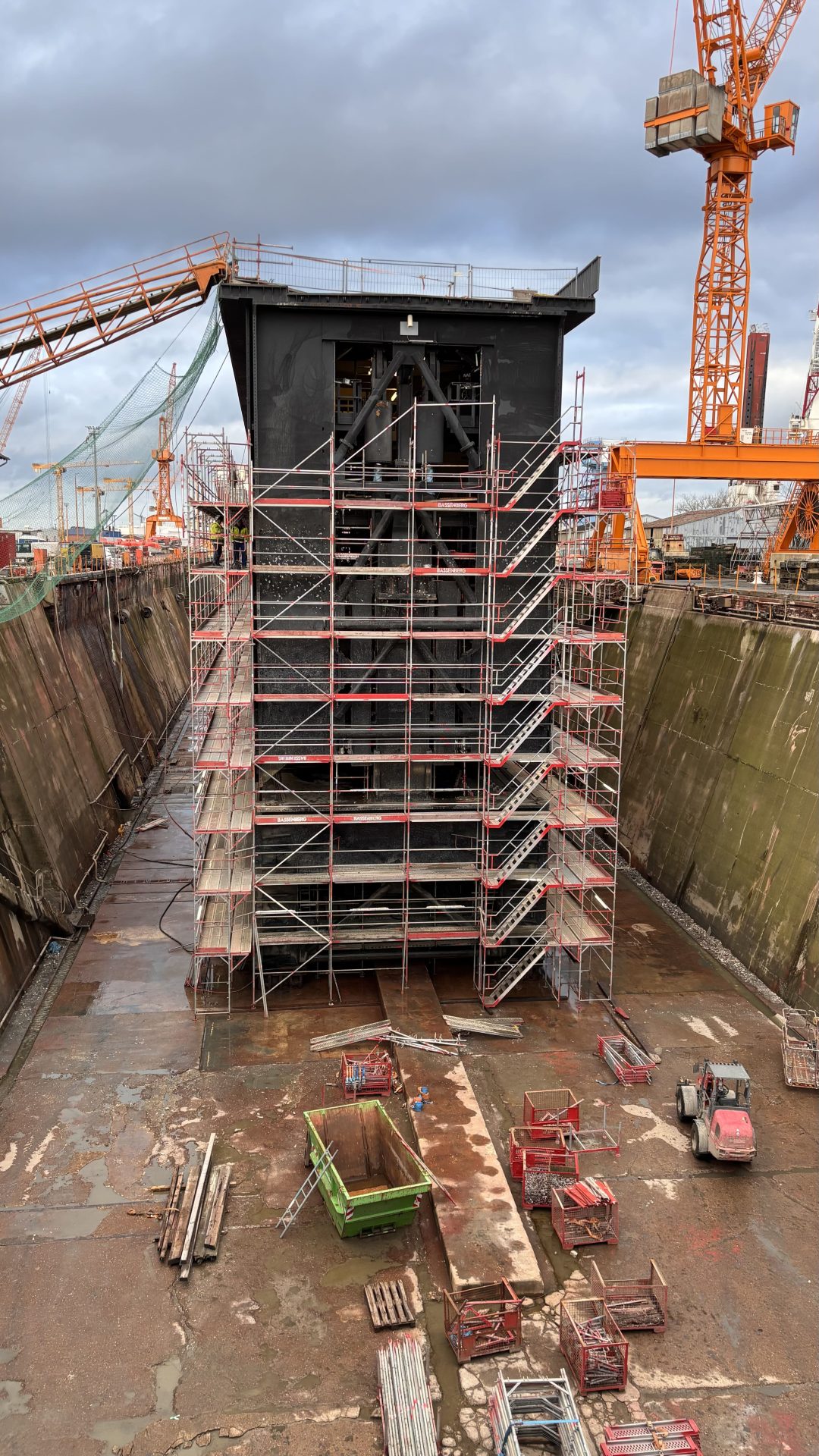 A large ship section under construction in a dry dock, surrounded by scaffolding and cranes, with building materials and equipment on the ground.