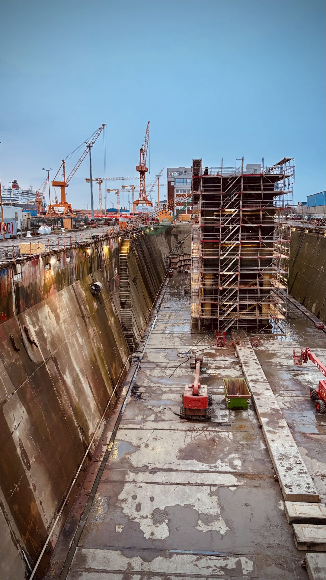 A dry dock with scaffolding around a large structure, cranes in the background, and construction equipment on the concrete floor.