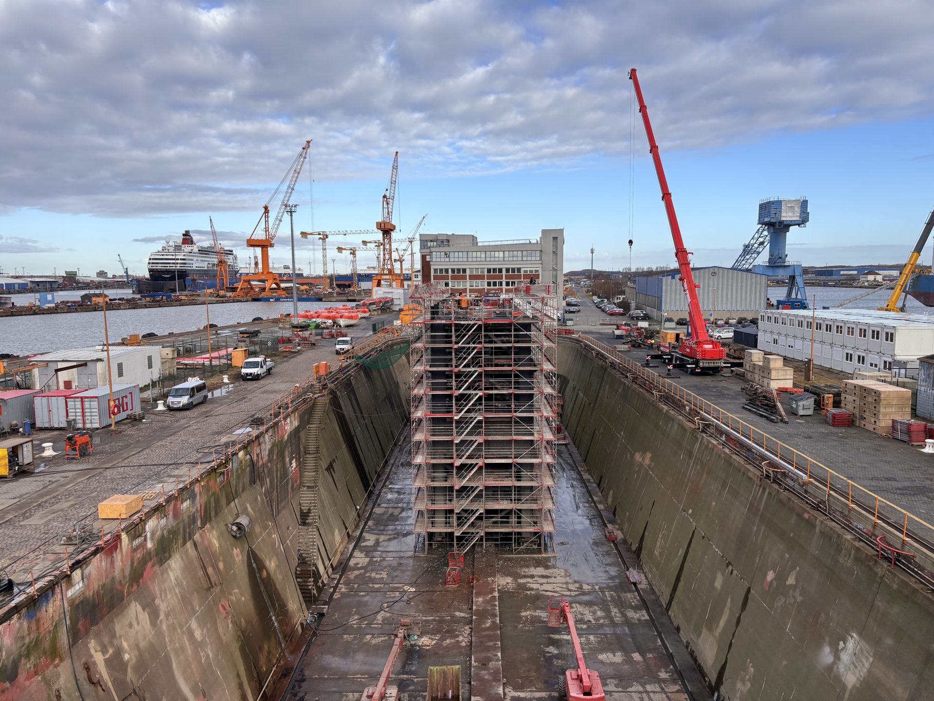 A large dry dock with scaffolding in the center is surrounded by cranes, vehicles, and industrial buildings under a partly cloudy sky.